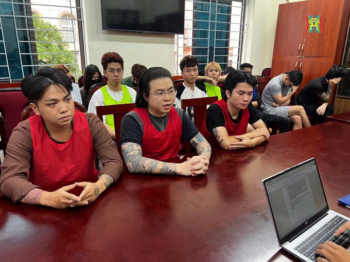 A group of young men wearing red and green vests sit around a conference table, appearing to be in a meeting or hearing, with a laptop in the foreground.