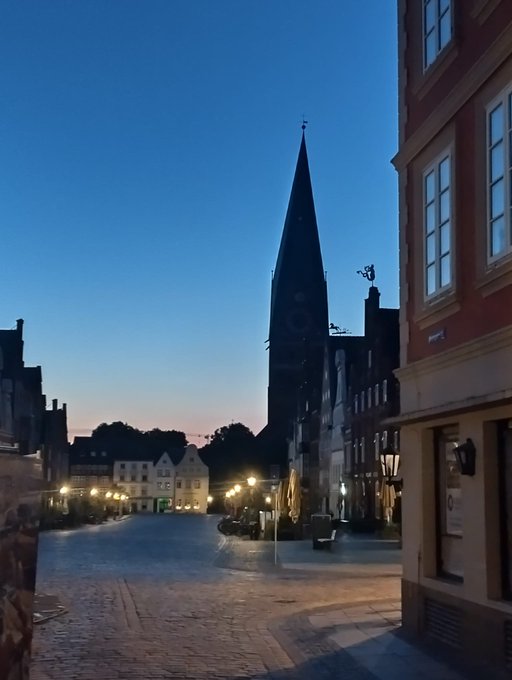 A twilight view of a European town square featuring a tall, pointed church spire, illuminated street lamps, and historic buildings.