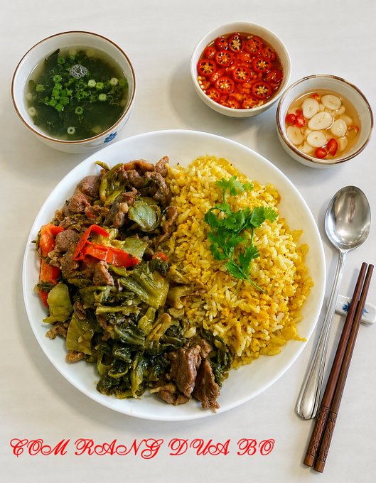A Vietnamese meal featuring a plate of yellow rice (Cơm Rang Dưa Bò) served with stir-fried beef and pickled mustard greens, accompanied by three small bowls of condiments including seaweed soup, sliced chilies, and pickled garlic, with a spoon and chopsticks on the side.