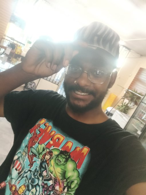 The image features a man wearing glasses, smiling for the camera while posing with his hand up to his face. The background of the photo shows a window, suggesting that they might be indoors or near an outdoor area like a patio. There are also several books visible in the scene, which could indicate a reading or study space nearby.