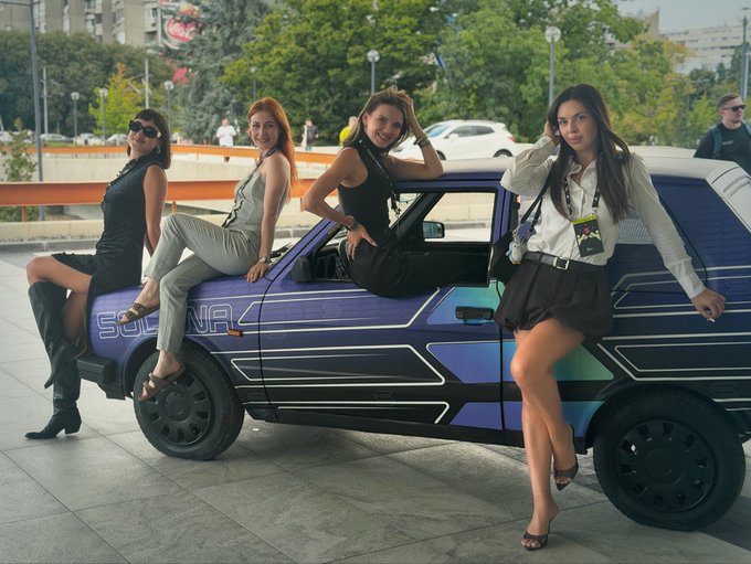 Photo of four women in fashionable outfits posing around a purple and blue futuristic-looking car labeled 'SOLANA' outdoors.