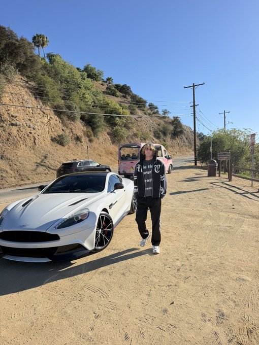 The image shows a person standing next to a white Aston Martin sports car on a dirt road in Los Angeles, with hills, power lines, and a pink van visible in the background. The caption aligns with the post's vibe of flexing the luxury vehicle under the sunny LA sky, while referencing crypto tickers like $CATE and $TA in hand.