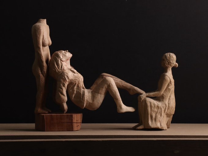 Three wooden sculptures—a standing faceless figure, a reclining woman, and a seated figure—rest against a dark background, their unfinished textures and intimate arrangement telling a quiet story of connection and solitude without words. The piece captures the narrative of three figures sharing a moment, embodying the post's invitation to convey an entire story through a single, evocative image.