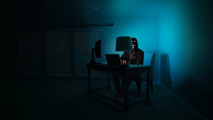 A hooded figure wearing a Guy Fawkes mask sits at a desk in a dark room, illuminated by a cyan light, symbolizing the hacker responsible for the Maya Protocol exploit. The image visually represents the cybercrime mentioned in the post, where a vulnerability allowed for the theft of millions in assets.