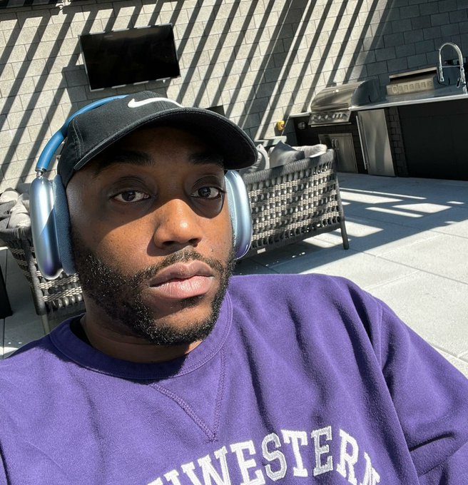 The image features a man wearing a purple sweatshirt with "Westester" written across the front, sitting outdoors with headphones on his head. The man appears to be smiling for the camera while wearing the headphones. There are also two TVs visible in the background, one of which is mounted on the wall and another placed on a table.