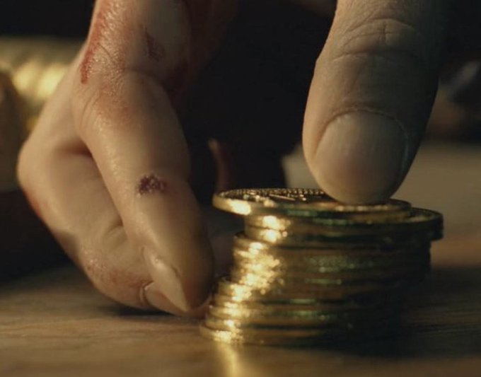A close-up cinematic shot shows a person's hand stacking gold coins on a wooden surface, with visible scratches on the fingers.