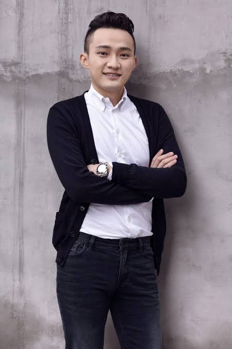 A portrait of a smiling East Asian man with short dark hair, wearing a white button-up shirt under a black cardigan, with his arms crossed in front of a textured gray wall.