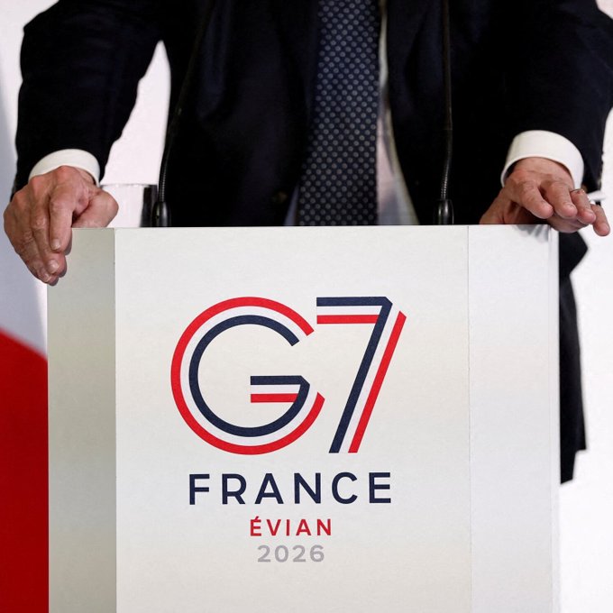 The image shows a man standing behind a podium, with his hands placed on top of it. The podium displays the text "THE G7 IS MEETING IN FRANCE RIGHT NOW AND THEY ARE TALKING BEHIND CLOSED DOORS Confidential sessions where they decide who gets rich in the new paradigm and who gets left holding fiat bags." This suggests that there is a significant event taking place, possibly related to cryptocurrency or financial matters. The man's position behind the podium indicates his involvement in the discussion or presentation at this gathering.