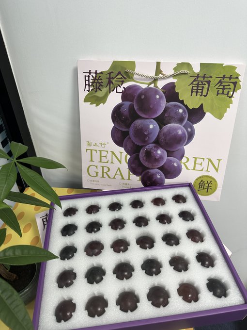 A purple gift box contains individually arranged dark purple grapes in foam padding, displayed in front of a card labeled 'TENGU GRAPES' with Japanese characters and an illustration of grapes.