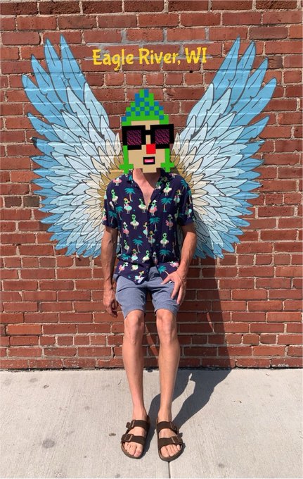 A person standing in front of a brick wall mural of blue angel wings, with their face covered by a pixel art character and the text "Eagle River, WI" displayed above.