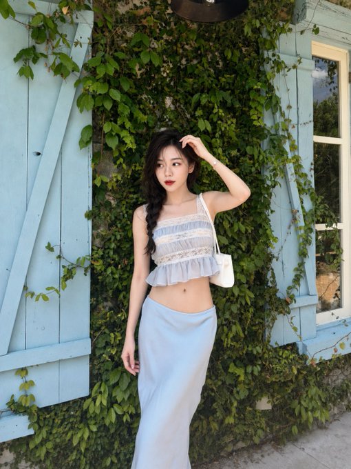 A young woman with long dark hair posing in a light blue two-piece outfit against a wall covered in green ivy and blue wooden shutters.