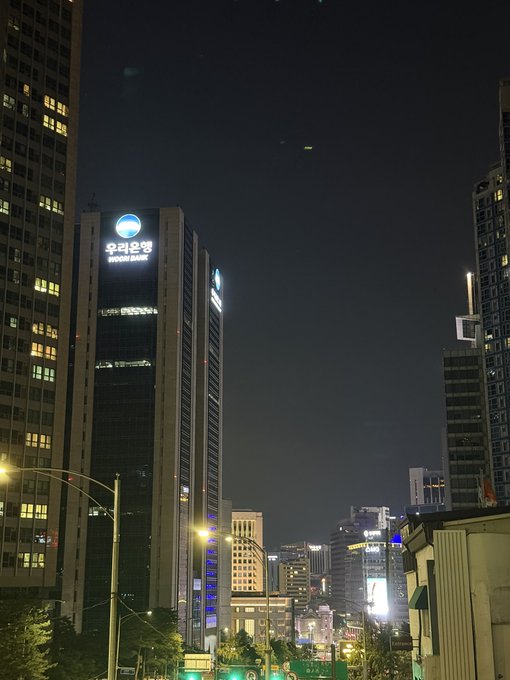 A nighttime cityscape of Seoul features the illuminated Woori Bank headquarters, its glowing blue logo and "우리은행" Korean signage standing out among the surrounding skyscrapers, capturing the late-hour office activity referenced in the post.