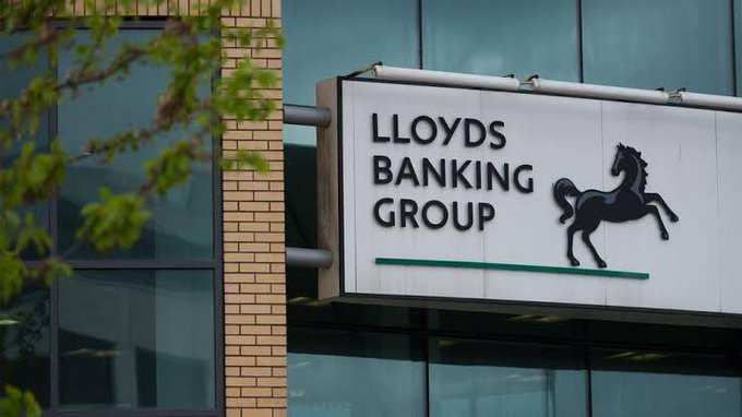 A photograph of a building sign reading "LLOYDS BANKING GROUP" with the company's signature black horse logo, mounted on a modern glass building with a tree branch in the foreground.
