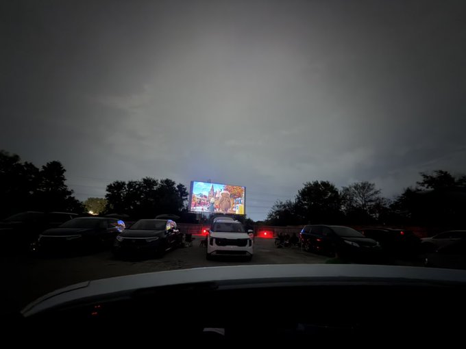 A nighttime view from inside a car at a drive-in movie theater, showing parked vehicles facing a brightly lit outdoor screen under a cloudy sky.