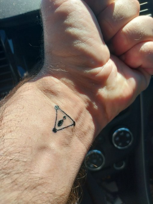 A close-up of a wrist inside a car shows a small black triangular tattoo or drawing containing an eye-like symbol.