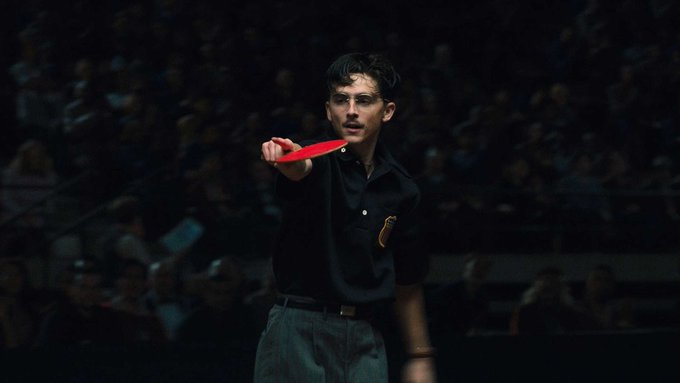 A young man in a dark shirt holds up a red object while standing before a dimly lit crowd, his intense expression suggesting focus and urgency—mirroring the post's message about resisting comparison and running your own race rather than feeling pressured by others' timelines.
