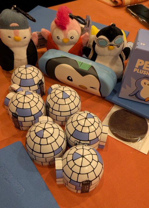 A display of plush toys including three penguin-style figures and a blue whale-themed pencil case, alongside five igloo-shaped containers, with a partially visible 'PE PLUS' sign.