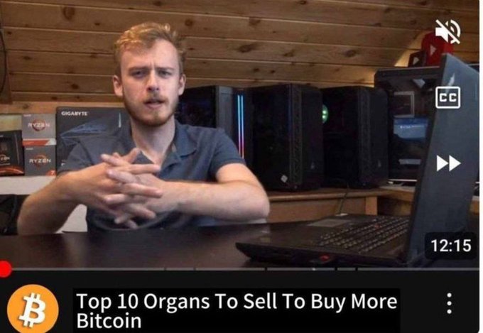 A young man with reddish-blonde hair sits at a desk with computer equipment and a laptop behind him, appearing to record a video with the timestamp 12:15 visible. The YouTube video is humorously titled "Top 10 Organs To Sell To Buy More Bitcoin," with a Bitcoin logo, poking fun at the desperation of crypto enthusiasts trying to buy the dip at any cost, as referenced in the accompanying post.