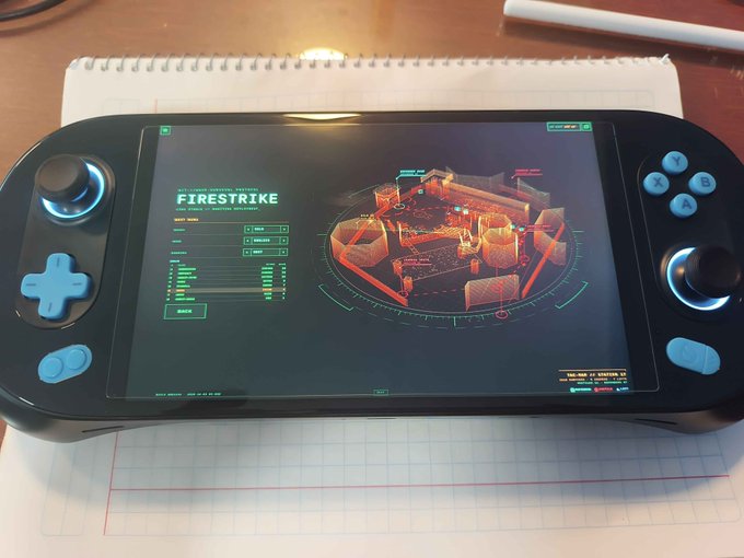 A black handheld gaming console rests on a spiral notebook, its screen displaying a 'FIRESTRIKE' menu with a tactical map interface.