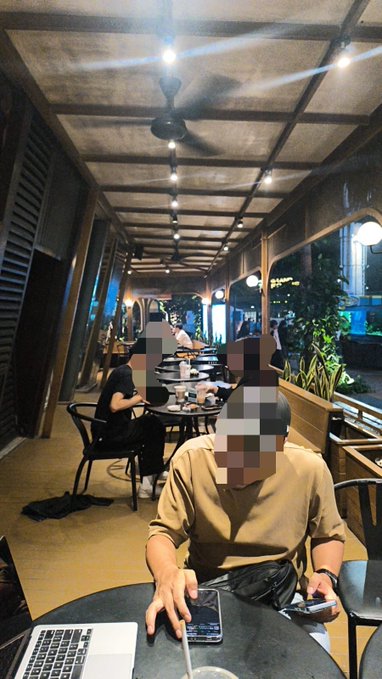 Two individuals sit at a table in a dimly lit, open-air restaurant with industrial ceiling beams and ceiling fans, their faces blurred, with one person in a tan shirt holding a smartphone next to an open laptop. The casual, on-site meeting setup aligns with the post's narrative of discussing the development of the DegenHunterApp and comparing it to ponyin.id/SIA.
