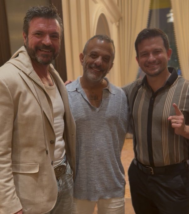Three men posing together for a photo, smiling, with the man on the left wearing a beige suit and the man in the center wearing a blue shirt.