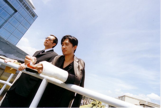 Two men stand on a balcony overlooking a harbor, one with a bandaged arm, in a scene from the Hong Kong film *Infernal Affairs* (无间道). The image pairs with the post's metaphor of perpetual hesitation—waiting for lower prices like waiting for "tomorrow"—echoing Chen Wing-yan's lament about never seizing the moment while it remains.