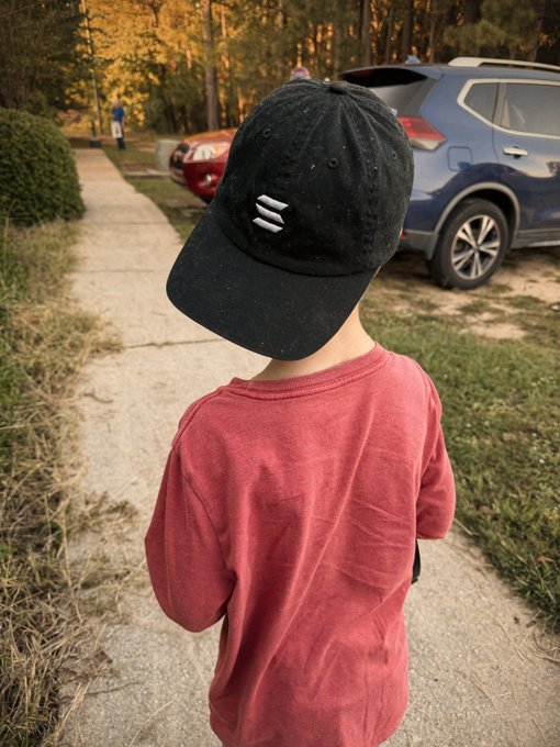 A young child viewed from behind wears a black cap featuring three stacked white horizontal lines and a faded red t-shirt while standing on a sidewalk near parked cars. The three-line logo on the hat represents the Solana blockchain symbol, tying into the "Solana Kids" branding of the post.