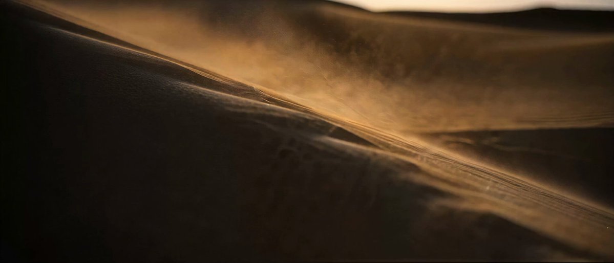 The image depicts a desert scene with sand blowing away. In the background, there are a few people standing near the dunes, who appear to be walking through or playing around them. The photo focuses more on the motion of the sand being blown by the wind and captures its movement, while also showing the presence of individuals in the desert setting.