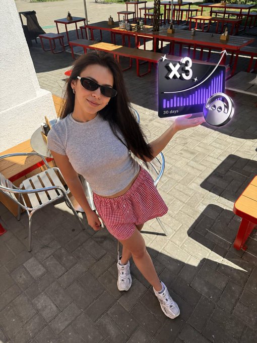 A woman in sunglasses, a gray tee, and a red gingham skirt poses outdoors near outdoor café seating while holding up an animated graphic displaying "x3" with "30 days" labeled and an upward growth chart, visually representing the promised triple yield over 30 days mentioned in the caption about activating Tangem Yield Mode through Aave.