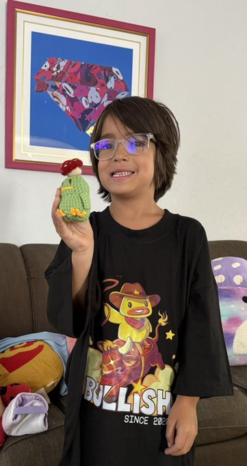 A smiling child wearing glasses and a black Bullish t-shirt holds up a small crocheted green cactus figure, with a framed artwork visible on the wall behind them.