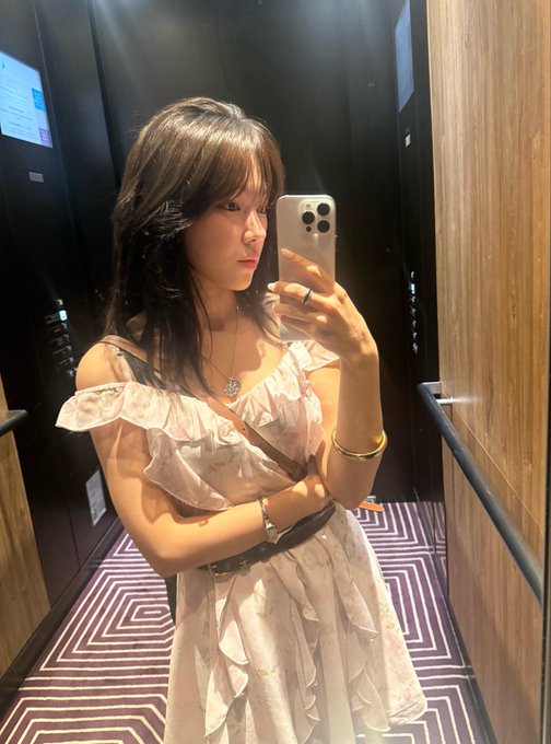A young woman with long dark hair takes a mirror selfie in an elevator hallway, wearing a ruffled white dress and gold jewelry while holding up an iPhone with a triple-camera setup. The sleek, modern interior with geometric patterned carpet and wood-paneled walls contrasts with the financial jargon in the caption, suggesting a moment of distraction or daydreaming about trading assets like PRL (Pearl) amid her stylish outing.