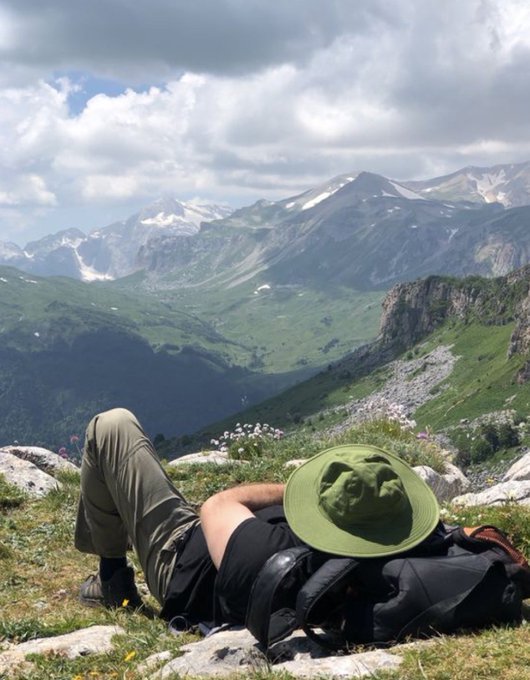 A hiker wearing a green wide-brimmed hat and olive pants lies on grassy alpine terrain, gazing out at snow-capped mountains under a partly cloudy sky.