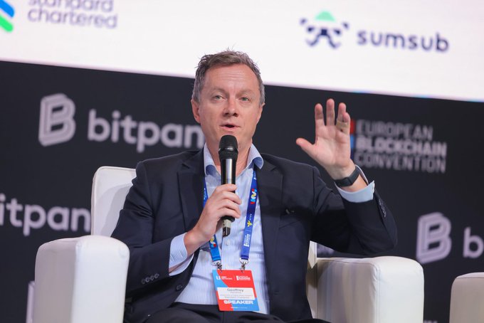 A man in a suit speaking into a microphone at the European Blockchain Convention, with 'standard chartered', 'sumsub', and 'bitpanda' logos visible in the background.