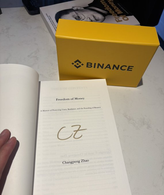 A book titled 'Freedom of Money' by Changpeng Zhao sits in front of a yellow 'BINANCE' promotional box.