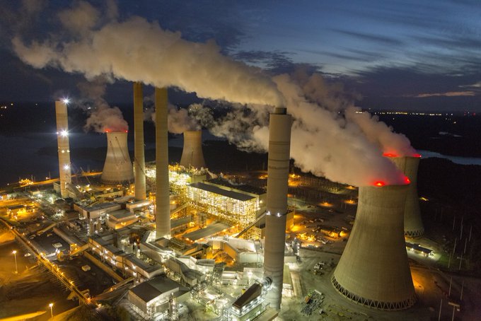 An aerial nighttime view of a large industrial power plant with illuminated cooling towers emitting plumes of white smoke into the dark sky.