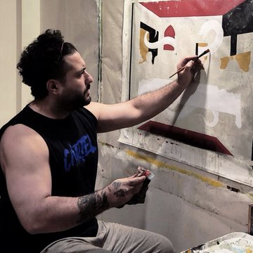 A tattooed artist in a black tank top sits working on an abstract painting with bold black, white, red, and yellow shapes mounted on a wall.