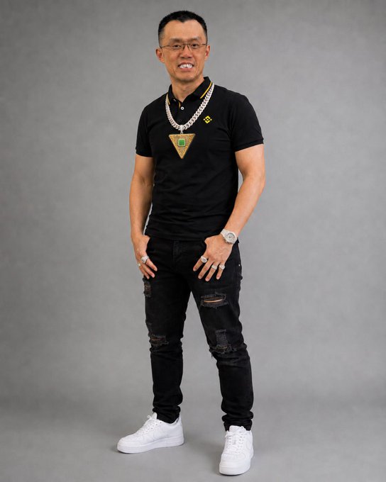 A full-body portrait of an East Asian man wearing glasses, a black polo shirt, black ripped jeans, white sneakers, and multiple thick silver chains, posing against a grey studio background.