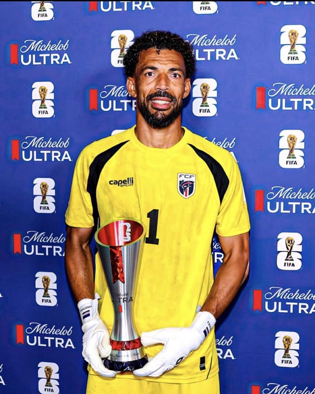 The image features a man wearing a yellow soccer uniform, holding a trophy with both hands while standing against a blue backdrop that includes logos for "Michelin Ultra" and "Capellia". The man appears to be posing for a photo or attending an event related to soccer.