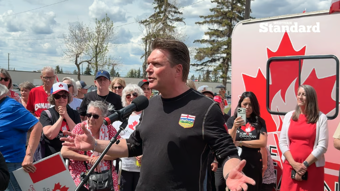 A man in a black shirt with a "Canada" patch speaks at an outdoor rally, with a crowd holding Canadian flags and a sign reading "Niagara Standard" behind him. The image relates to the post's criticism about a politician delaying financial disclosure reporting.