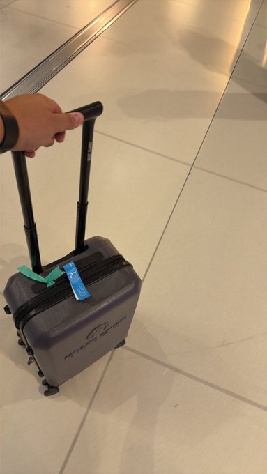 A hand grips the extended handle of a small gray carry-on suitcase (with a distinctive blue luggage tag) rolling across a tiled floor, ready for the road. The image perfectly complements the caption about hitting the road and trading from anywhere.