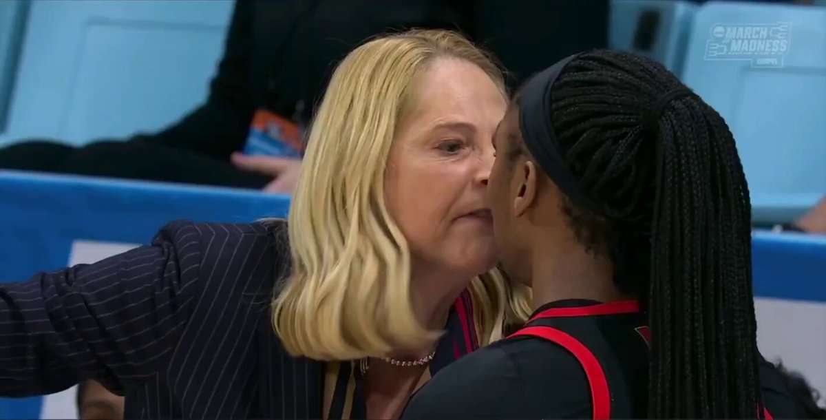The image shows two women standing next to each other with their mouths close together, possibly kissing or having a conversation. The background of the photo features a blue wall, which could be part of an event or gathering taking place at a stadium. There is also a person sitting in the background who appears to be watching something on a screen.