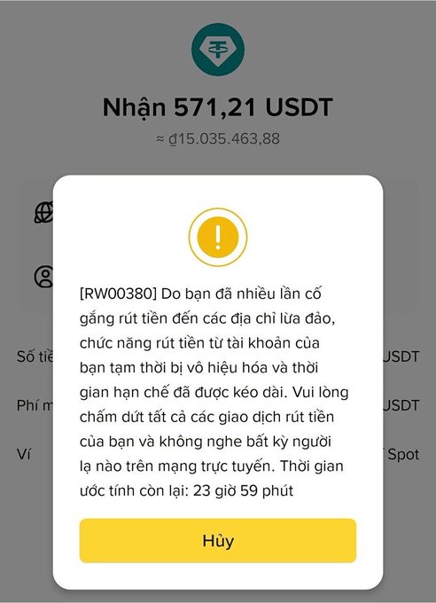 A Binance withdrawal screen showing a pending receipt of 571.21 USDT (≈₫15,035,463.88) is blocked by a warning popup labeled [RW00380], stating that the user has repeatedly attempted to withdraw to a fraudulent address, so the withdrawal function has been temporarily suspended for an estimated 23 hours and 59 minutes, with a "Huỷ" (Cancel) button to dismiss it. This illustrates the post's point that Binance's security system automatically detects scam addresses and prevents withdrawals to protect users from being tricked by clipboard-hijacking hacks.