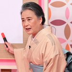 The image shows an older woman wearing a traditional pink kimono with a white obi, holding up what appears to be a red-handled object while speaking. The accompanying post humorously compares the scene to a "hardworking CEO riding in business class, laptop open," suggesting the woman's poised, on-the-go demeanor resembles a busy executive mid-travel.