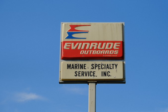 A vintage "Evinrude Outboards" and "Marine Specialty Service, Inc." sign stands against a clear blue sky, showing a weathered, nostalgic aesthetic. This retro signage evokes themes of classic Americana and bygone eras, complementing the "Catch & Release" NFT artwork mentioned in the post by hinting at themes of fishing and maritime culture.