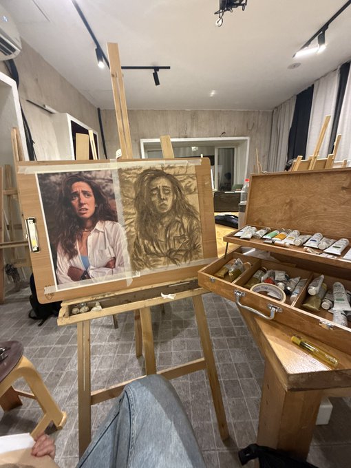 An art studio scene showing an easel with a portrait painting in progress alongside its reference photo of a woman, next to a wooden box of oil paints.