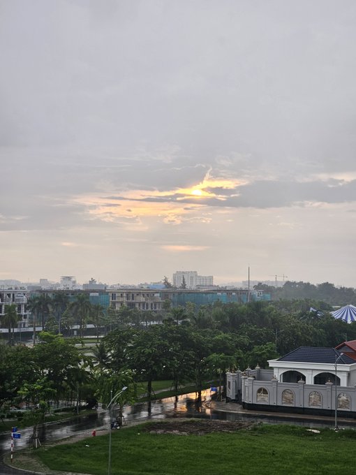 A landscape photograph taken at sunset showing a residential area with trees, buildings, and a gateway, with the sun breaking through cloudy skies.