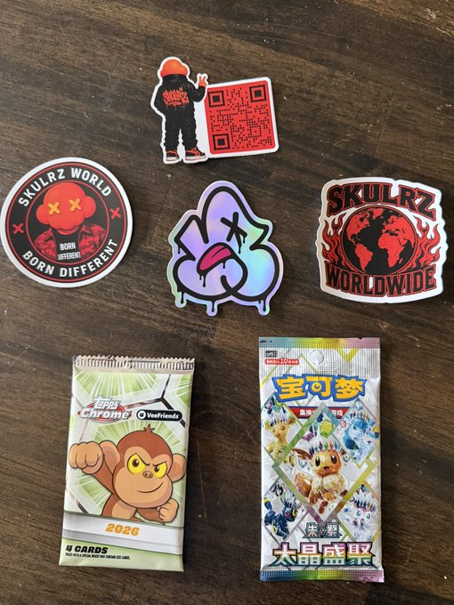 Various 'Skullrz World' and 'Skullrz Worldwide' stickers are arranged on a wooden surface alongside two Pokemon card packs - one featuring Chimchar and another Japanese Pokemon pack.