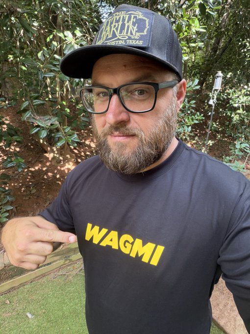 A selfie of a bearded man wearing glasses and a black 'WAGMI' t-shirt, pointing at the text while wearing a black 'ARTIFAX' cap from Austin, Texas, outdoors near greenery.
