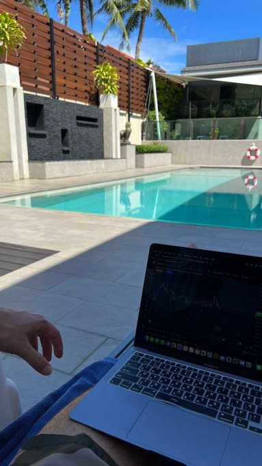 A first-person view of someone sitting by a swimming pool with a MacBook displaying financial charts on their lap, with palm trees and a modern building visible in the background.