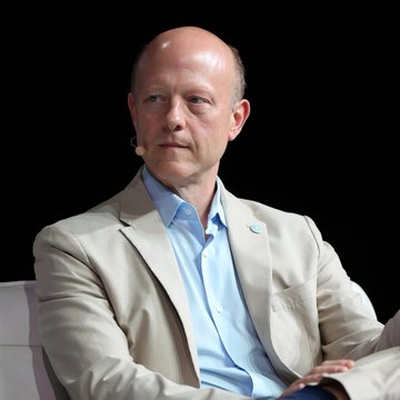 The image features a bald man wearing a suit, sitting down and looking off into the distance. The background of the photo is black, which contrasts with the white shirt he's wearing. There are no other people or objects visible in the picture. The caption provided seems unrelated to the main subject matter - it reads "BREAKING Circle to enter Japanese corporate market with Nomura for instant foreign currency settlement - Nikkei." This could be a reference to an event, product, or service related to the Japanese corporate market and Nomura's services.